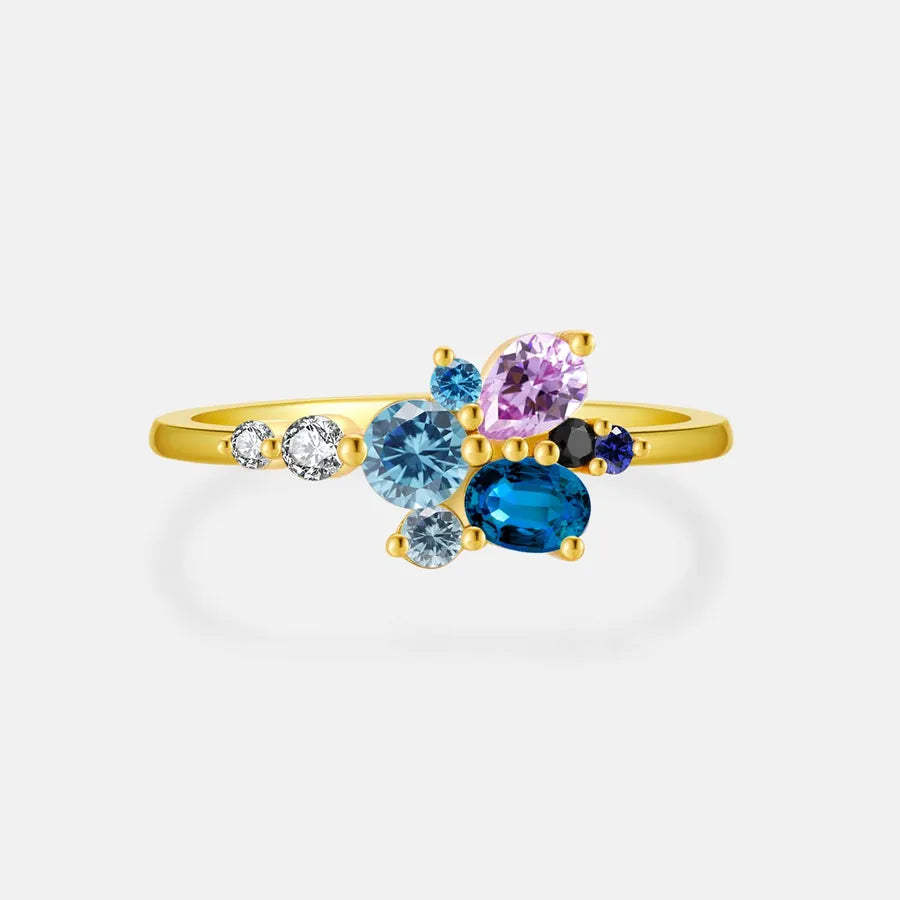 A gold ring featuring a cluster of colorful gemstones, including a large light blue oval stone, a larger dark blue oval stone, a lavender pear-cut stone, a small black round stone, and clear accent stones, set against a light gray background.