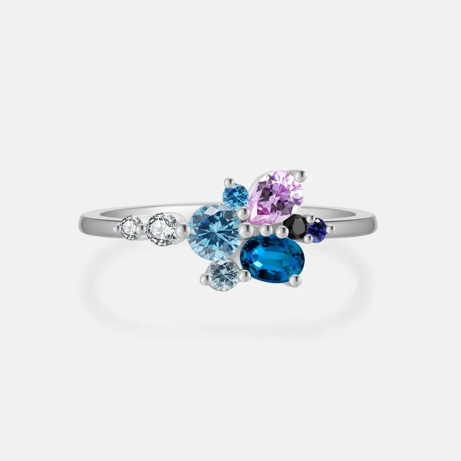 A silver ring featuring a cluster of colorful gemstones, including a large blue oval stone, a pink pear-cut stone, a light blue round stone, a small dark blue round stone, and a tiny clear accent stone, set against a light gray background.
