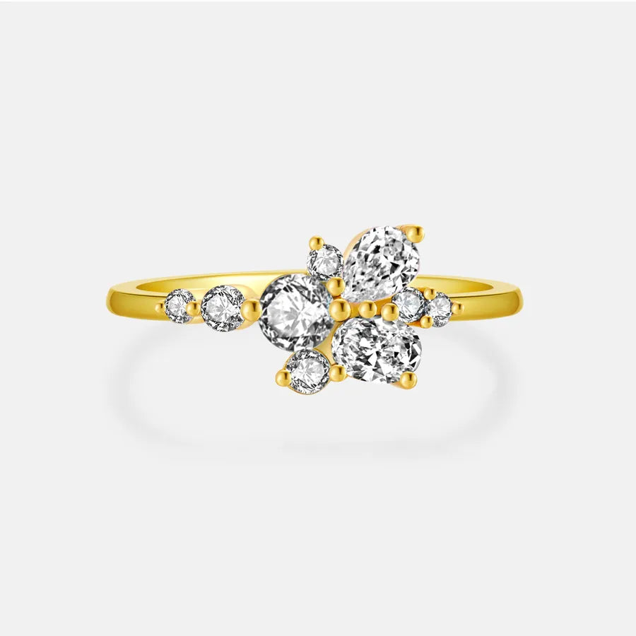 A yellow gold ring featuring a cluster of clear round-cut and oval-cut gemstones in a floral arrangement, set against a light gray background.