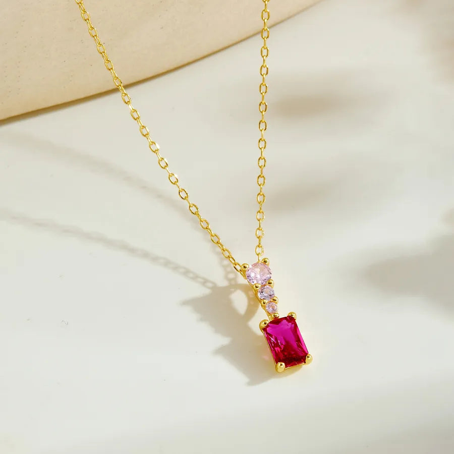 A gold chain necklace with a rectangular pink gemstone pendant, accented by a small pink heart-shaped stone and a round clear stone, displayed on a light beige surface.