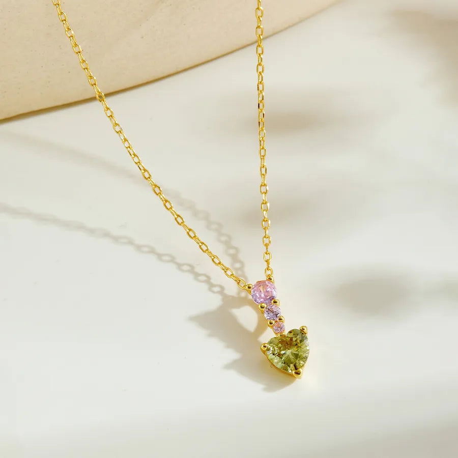 A gold chain necklace with a heart-shaped pendant featuring a light green heart, a small pink heart, and a tiny clear heart, displayed on a warm off-white surface.