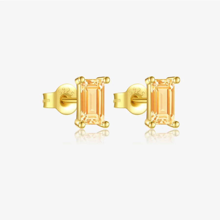 A pair of gold stud earrings with rectangular baguette-cut peach-colored gemstones, displayed on a clean white background with a soft reflection below.