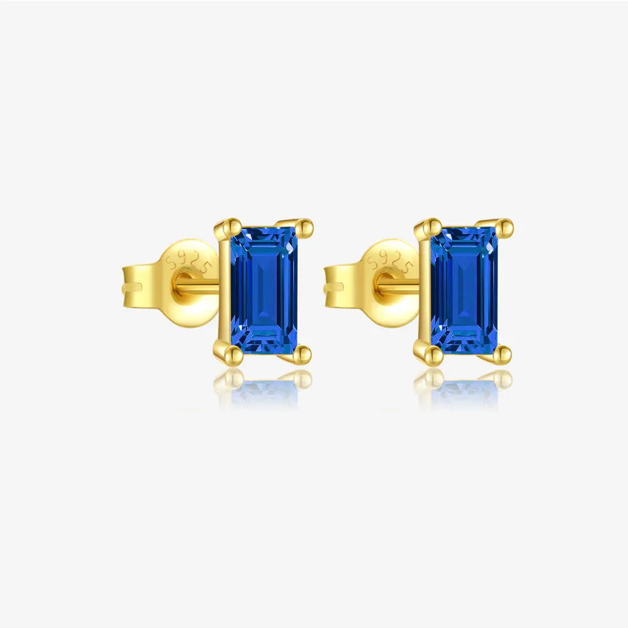 A pair of yellow gold stud earrings featuring rectangular blue baguette-cut gemstones in prong settings, shown against a light gray background with a soft reflection below.