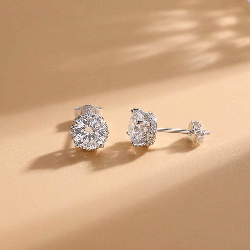 A pair of silver stud earrings with clear round-cut gemstones, shown on a warm beige background with soft leaf shadows.