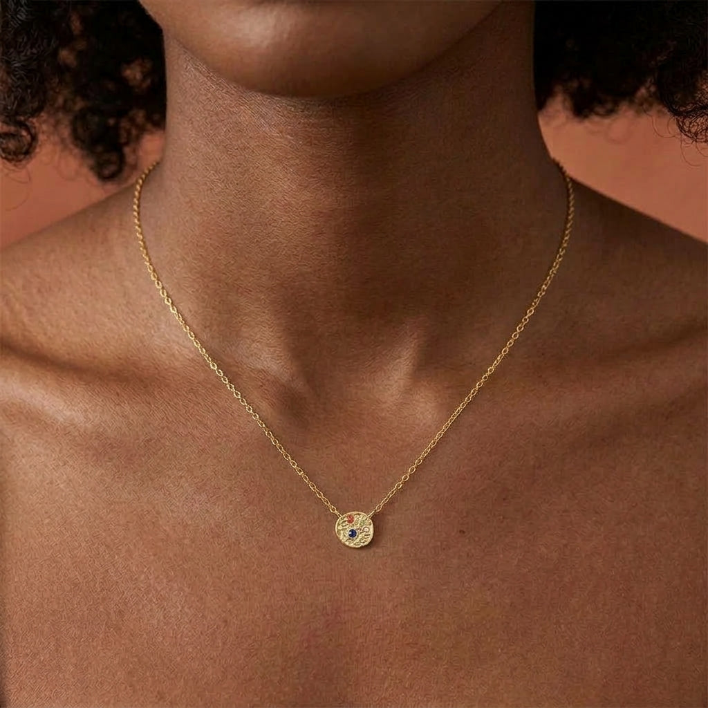 A close-up of a model's neck and chest wearing a delicate gold chain necklace with a small circular pendant featuring a blue stone.
