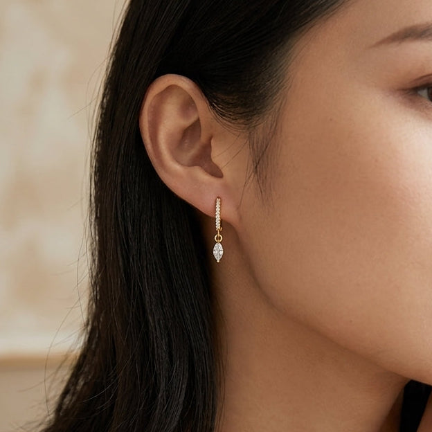 A close-up lifestyle shot of a woman with dark hair wearing delicate gold jewelry, including a small drop earring, a delicate necklace, and a bracelet, set against a soft, warm beige background.