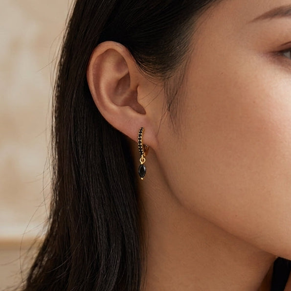 A close-up lifestyle shot of a woman with dark hair wearing delicate gold jewelry, including a small gold hoop earring with dark green stones, a gold necklace, and gold bracelets.