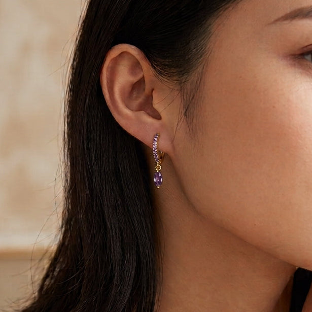 A close-up lifestyle shot of a woman with dark hair wearing a delicate gold necklace and matching gold bracelets, with a small gold drop earring featuring a purple gemstone accent.