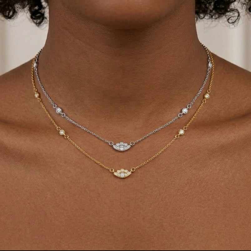 A close-up of a model wearing two delicate layered necklaces: a silver chain with a small oval clear stone pendant and a gold chain with a larger oval cream/beige stone pendant, both accented with tiny round stones.