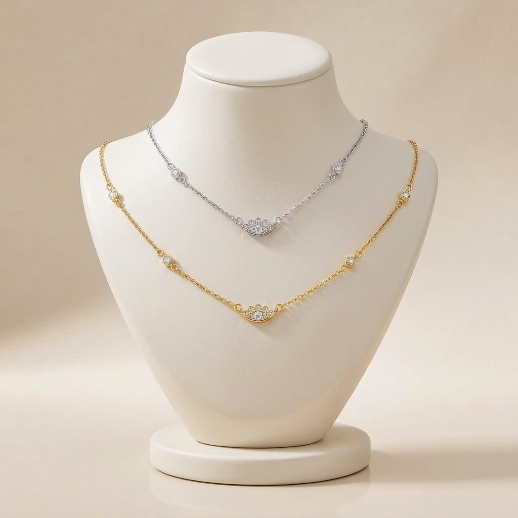 Two delicate necklace chains displayed on a white jewelry bust against a warm beige background. The lower chain is gold with small clear stone accents, and the upper chain is silver with a larger clear stone centerpiece.