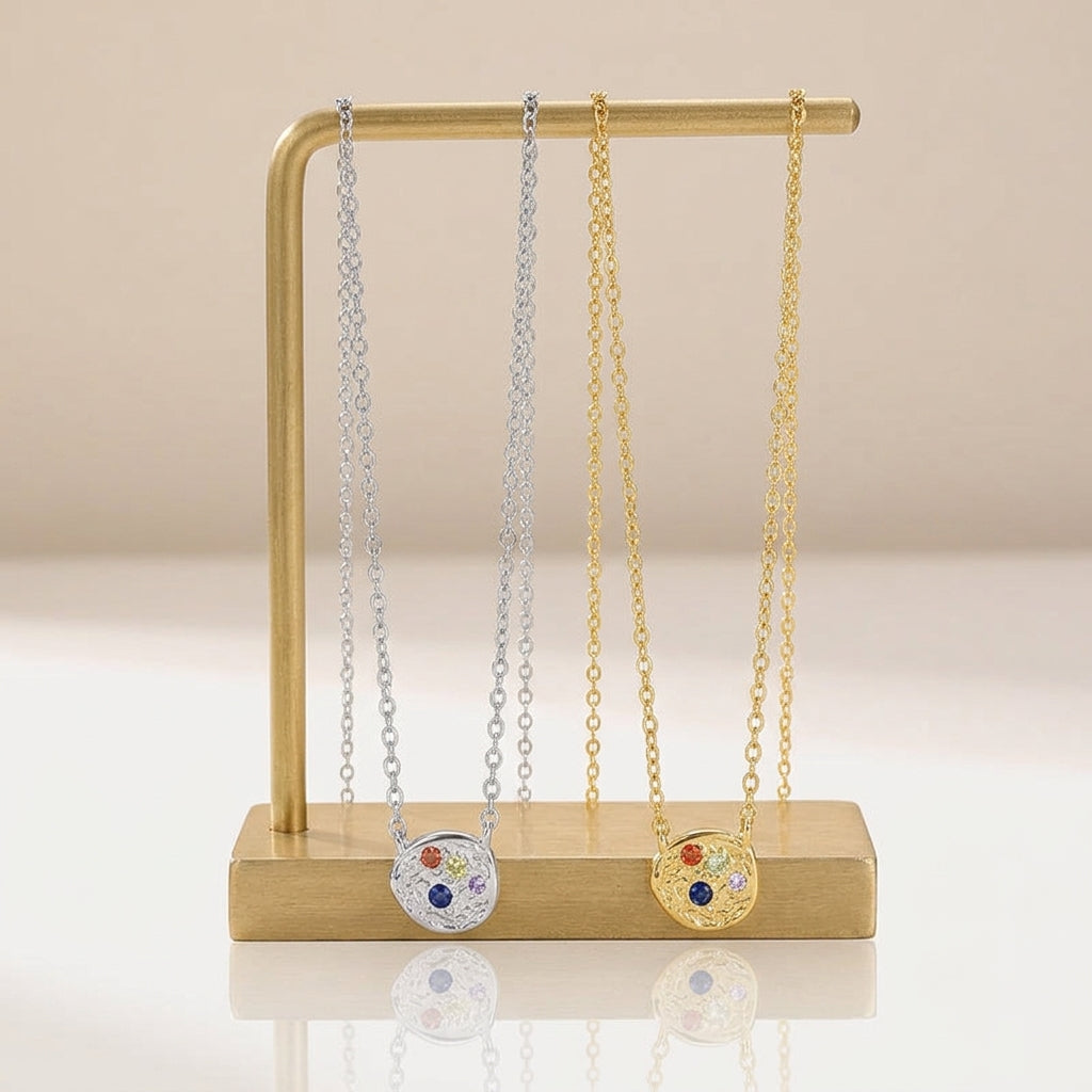 Two round pendant necklaces displayed on a gold stand, one in silver and one in gold, each featuring a colorful gemstone center on delicate chains against a warm beige background.