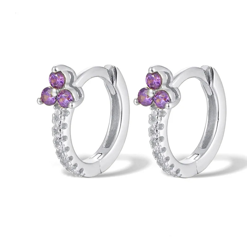 A pair of silver huggie hoop earrings with a small flower cluster of purple gemstones at the top and a row of clear cubic zirconia stones along the front, set against a plain white background.
