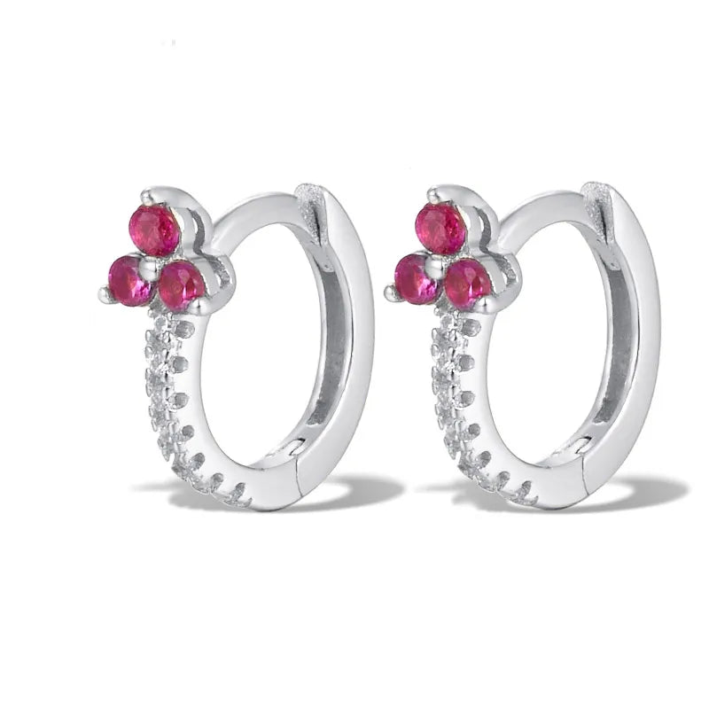 A pair of silver huggie hoop earrings decorated with three small pink gemstones on each earring, accented by a row of tiny clear stones along the front curve, set against a plain white background.