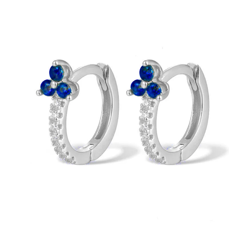 A pair of silver huggie hoop earrings featuring a small flower cluster of three deep blue stones at the top and a row of clear pavé stones along the lower curve, set against a plain white background.