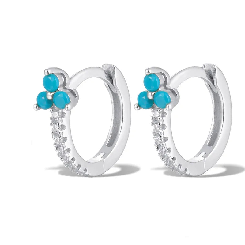 A pair of silver huggie hoop earrings featuring a small cluster of turquoise blue stones at the top and a row of clear cubic zirconia stones along the lower curve, set against a plain white background.