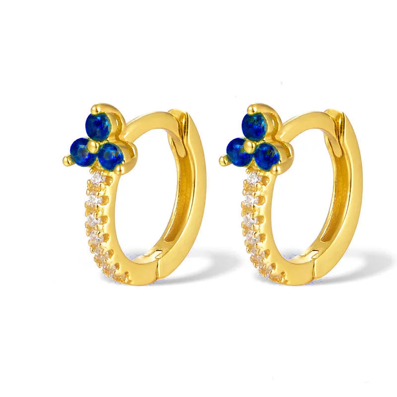 A pair of gold huggie hoop earrings featuring a small flower motif at the top with three deep blue stones and a curved row of small clear stones along the lower edge, set against a plain white background.