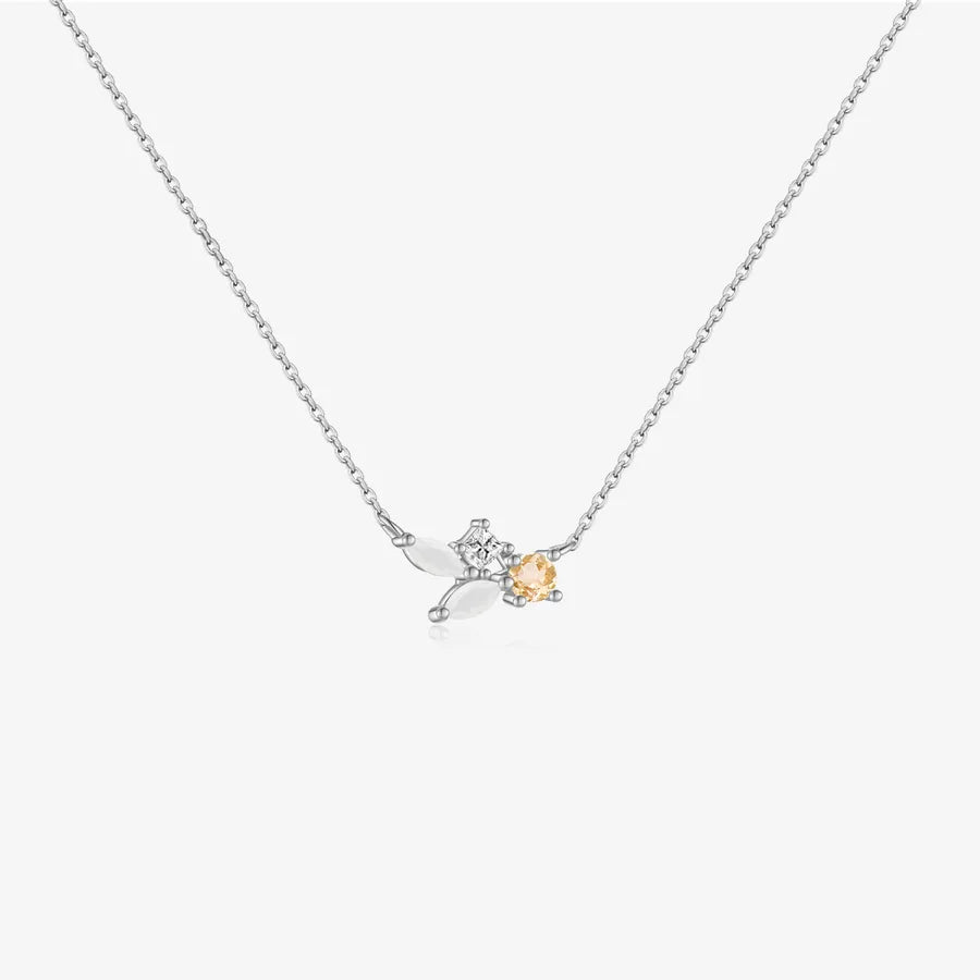 A delicate silver chain necklace featuring a small pendant with two silver leaves and a central clear crystal, accented by a small peach-colored heart-shaped gemstone, shown on a light gray background.