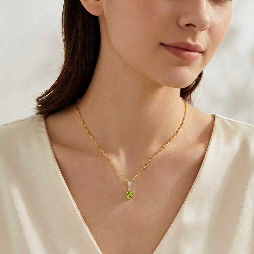 A close-up of a woman wearing a delicate gold chain necklace with a small green heart-shaped pendant, paired with a cream satin top.