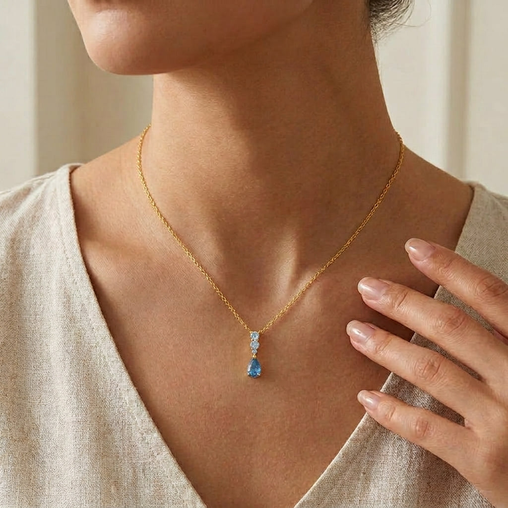 A close-up of a model wearing a delicate gold chain necklace with a small blue teardrop pendant and clear accent stones.