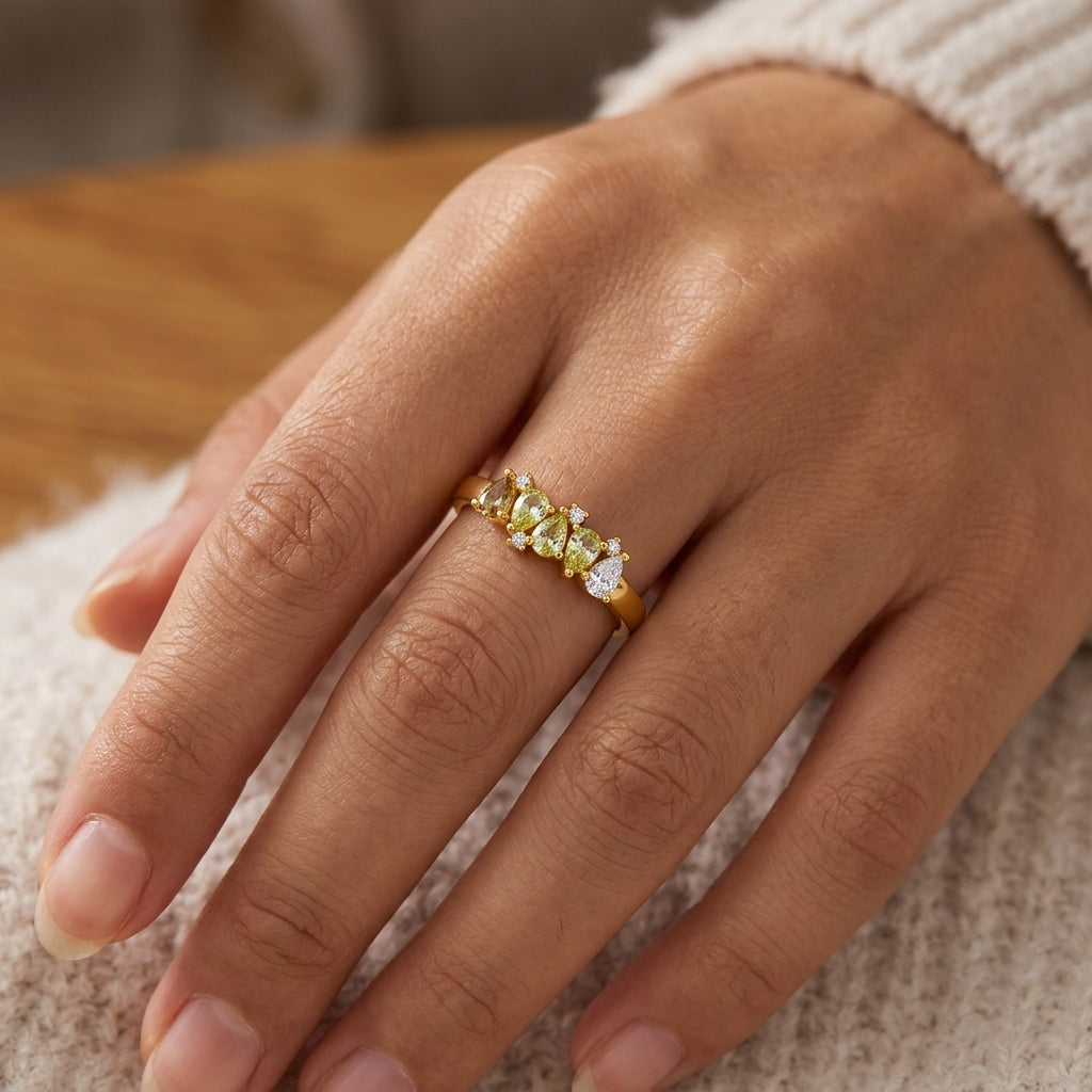 A close-up of a hand wearing a gold ring adorned with small yellow and green stones, resting on a soft cream knit surface.