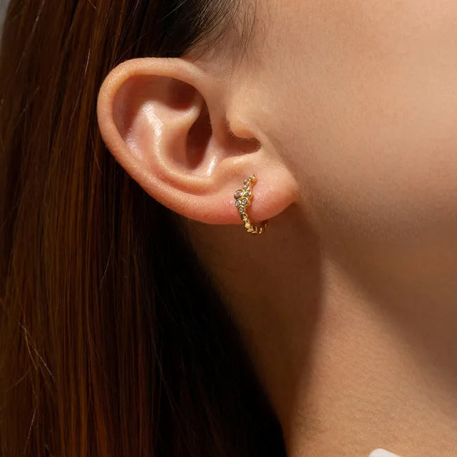 A close-up of a person's ear wearing a small gold huggie hoop earring adorned with tiny clear stones.