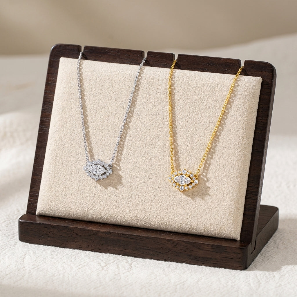 Two eye-shaped pendant necklaces displayed on a beige cushion inside a dark wooden stand. One necklace is silver-toned with a clear gemstone pendant, and the other is gold-toned with a matching clear gemstone pendant.