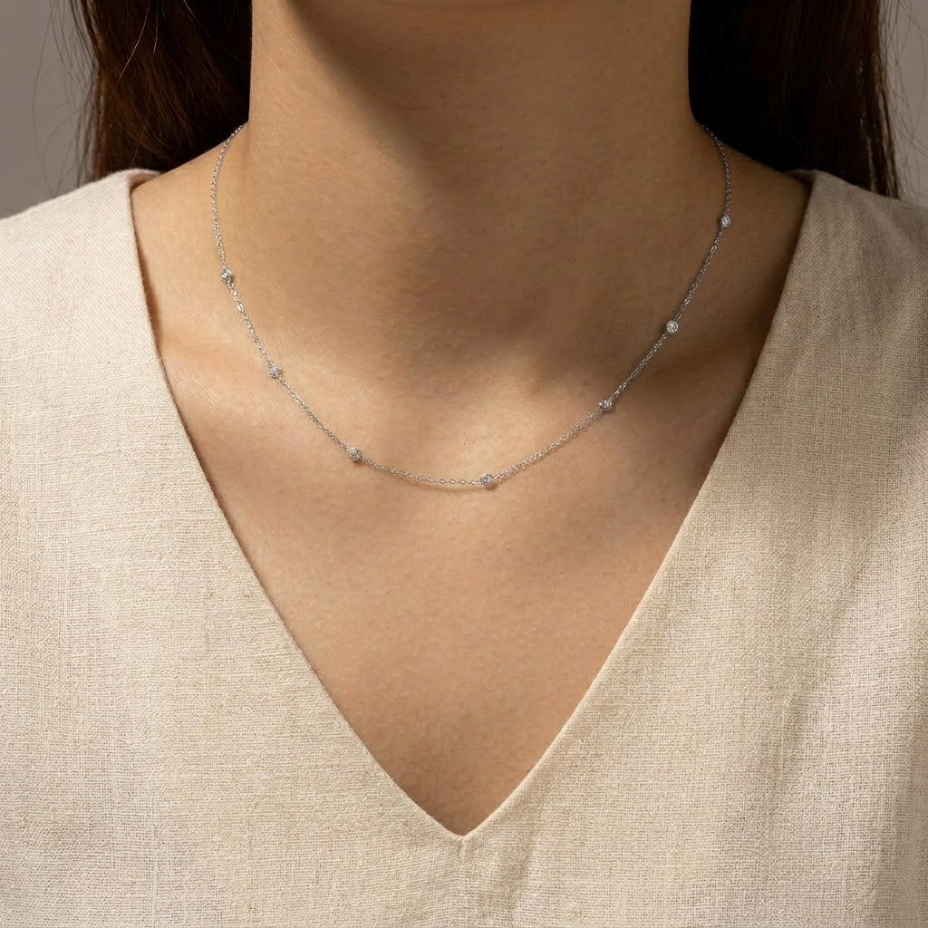 A delicate silver chain necklace with small, evenly spaced round clear stones, shown worn on a model's neck over a beige linen V-neck top.