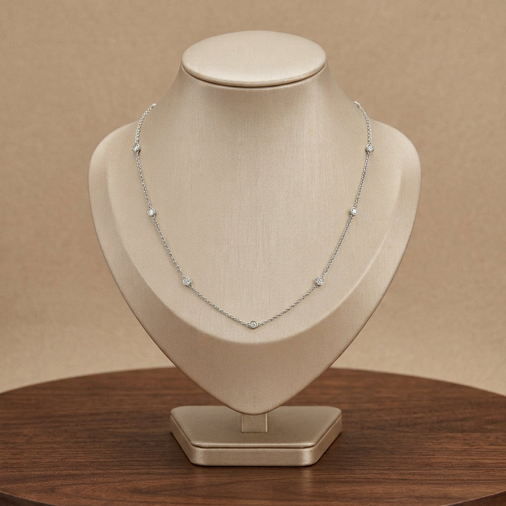A delicate silver necklace with small sparkling bead accents displayed on a beige jewelry bust stand, set against a warm beige background and a dark wood surface.