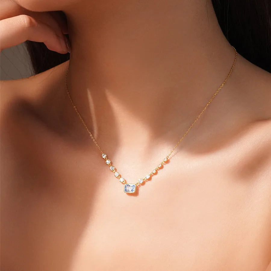 A delicate gold necklace with a small rectangular clear gemstone pendant, worn on a model's neck against warm sunlight.