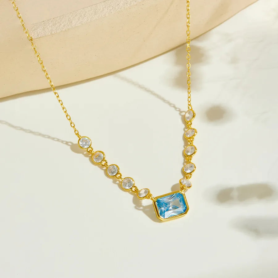 Gold chain necklace with a central rectangular light blue gemstone pendant, accented by small clear round stones along the chain, displayed on a light cream surface.