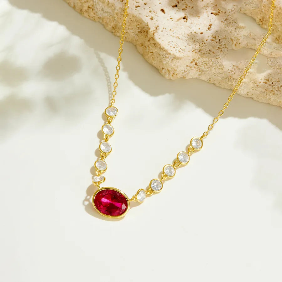 A gold chain necklace featuring a prominent oval-cut red gemstone pendant, accented by smaller clear round stones and tiny pearl-like beads, displayed on a light surface with a textured stone prop.