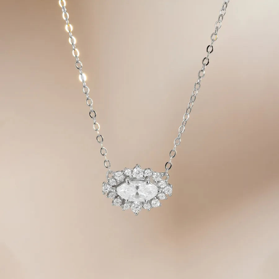A silver-toned necklace with a marquise-shaped clear gemstone surrounded by small round stones, suspended on a delicate chain against a soft beige background.