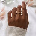 A close-up of a hand wearing a delicate gold ring with small white stones, styled with dried pink grass stems on a soft white fabric background. A perfume bottle and a folded scarf are softly blurred in the background.