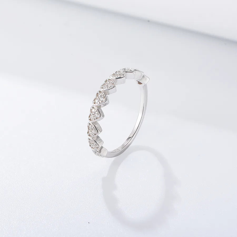 A silver band ring featuring a row of small, heart-shaped clear gemstones, photographed on a clean white surface with a soft reflection.
