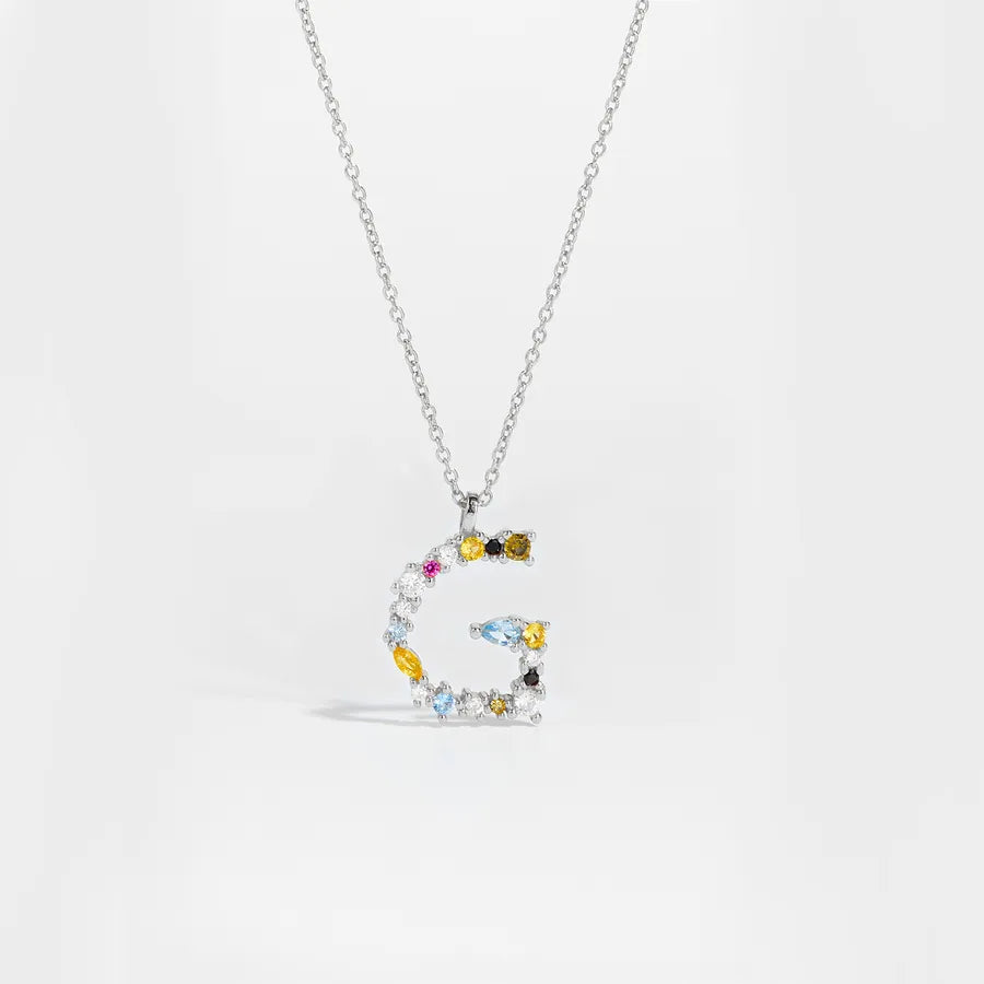 A silver letter 'G' pendant necklace adorned with multiple colorful gemstones, including yellow, light blue, dark brown, and pink, on a clean white background.