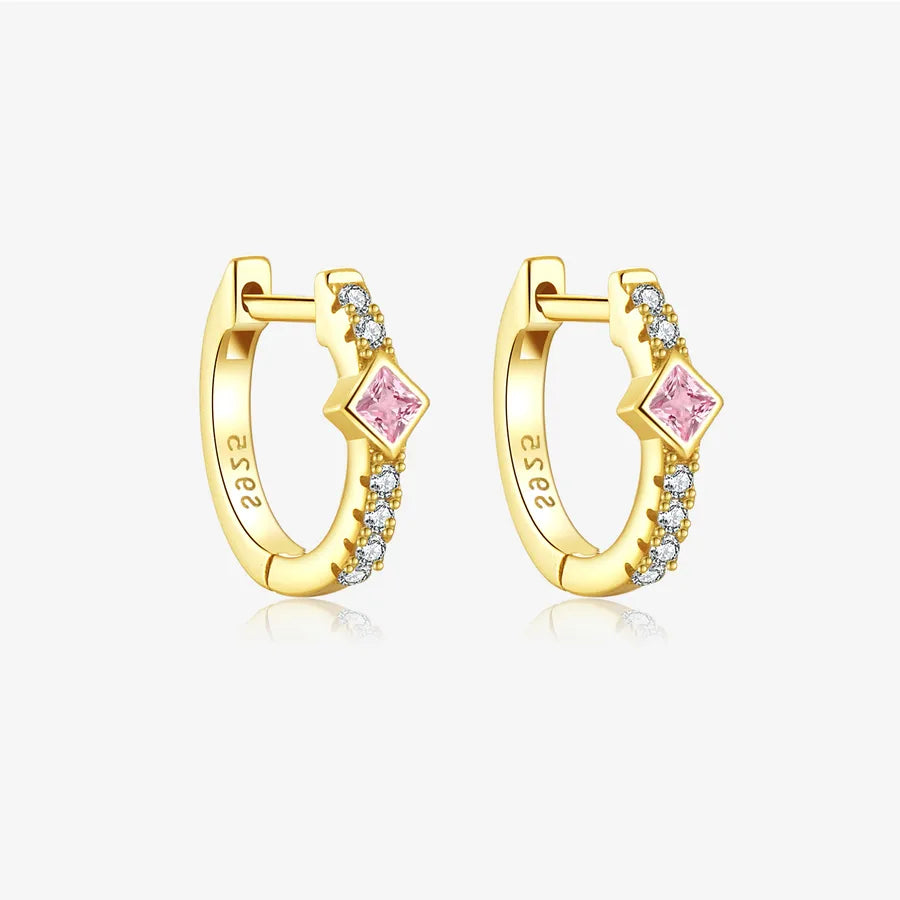 A pair of gold huggie hoop earrings with a central pink diamond-shaped stone and small clear stones along the band, shown on a light gray background.