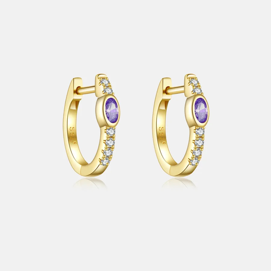 A pair of gold huggie hoop earrings, each featuring a round purple amethyst center stone and a row of small clear cubic zirconia stones along the outer curve, set against a light gray background.