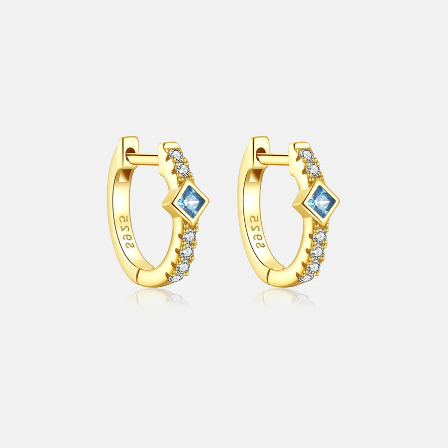 A pair of gold huggie hoop earrings with a central blue diamond-cut stone and small clear accent stones, shown on a light gray background.