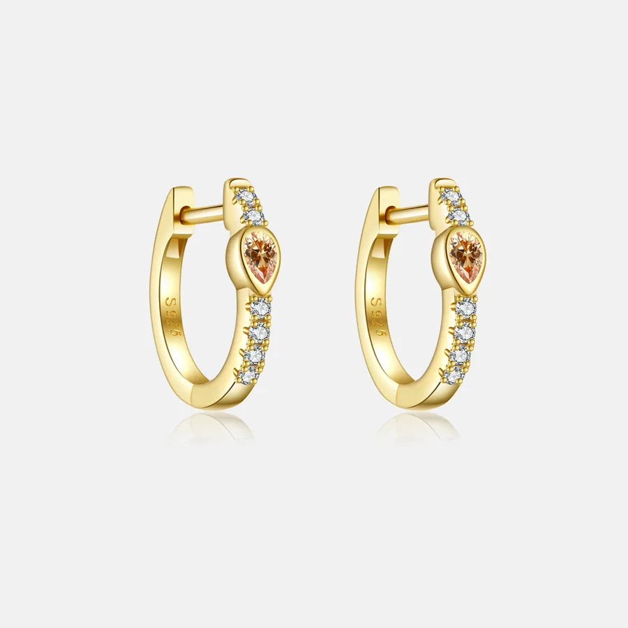 A pair of gold huggie hoop earrings with a central heart-shaped orange gemstone and small clear stones along the band, set against a light gray background.