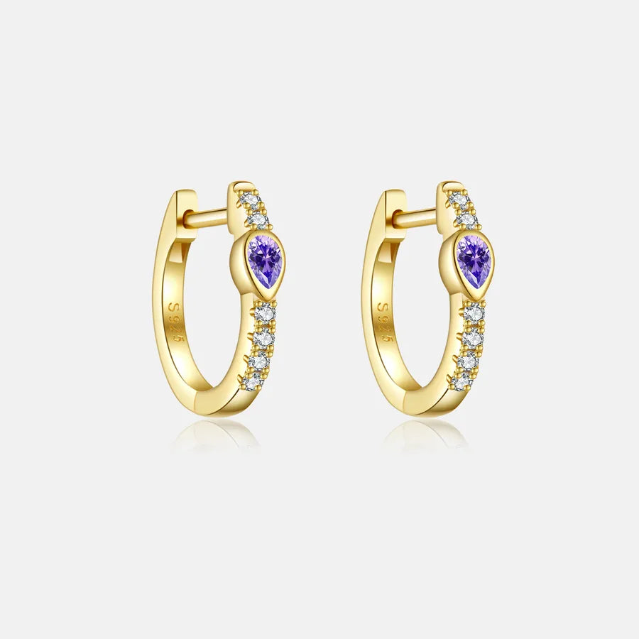 A pair of gold huggie hoop earrings with a central heart-shaped purple gemstone and small clear accent stones along the band, shown on a light gray background.