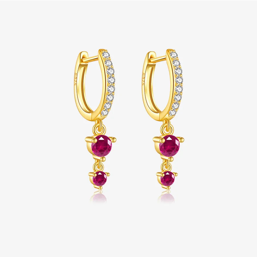 A pair of gold huggie hoop earrings with small clear stones along the front and three dangling round ruby gemstones, shown on a light gray background.