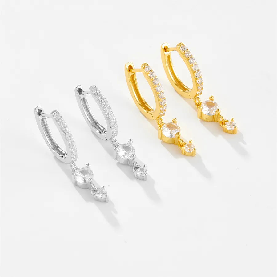 Two pairs of small hoop huggie earrings with dangling clear crystal drops, shown on a clean white background. One pair is silver-toned and the other is gold-toned, both adorned with small clear stones along the hoop.