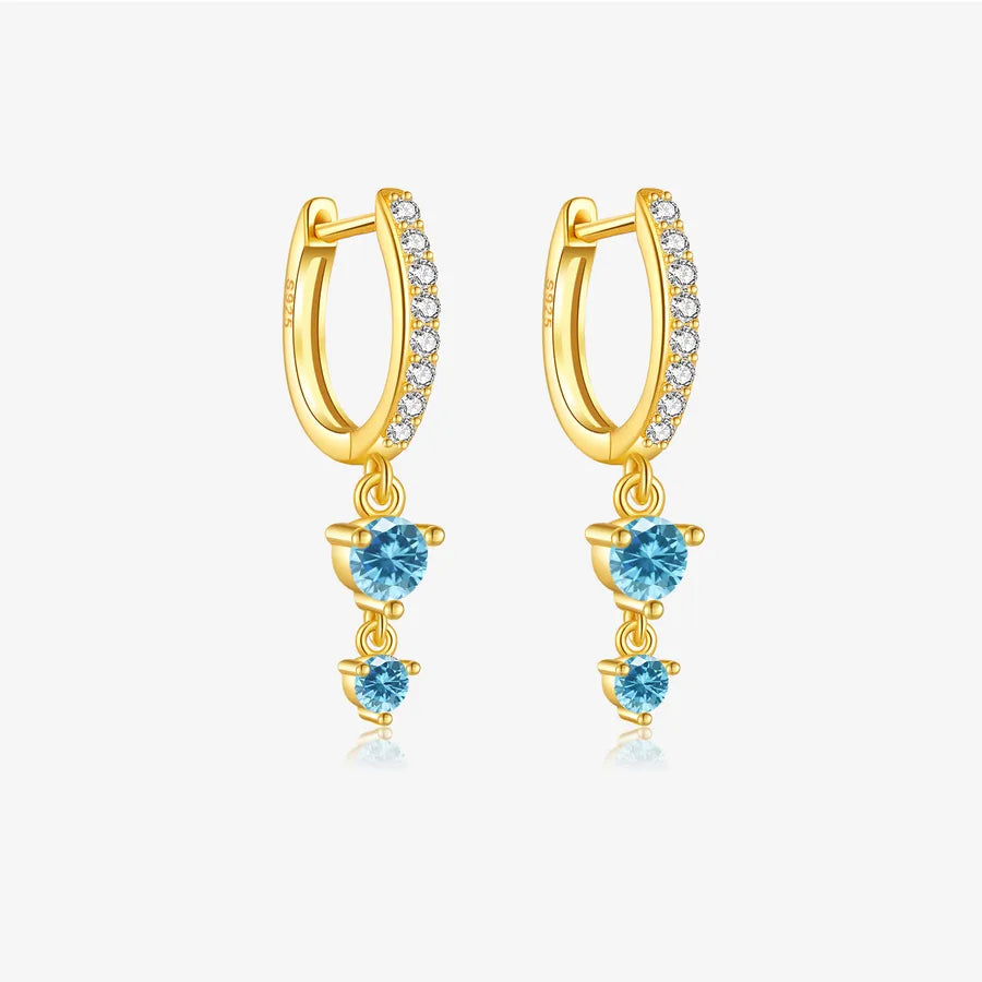 A pair of gold huggie hoop earrings with small clear stones along the front and three dangling light blue heart-shaped gemstones, shown on a light gray background.