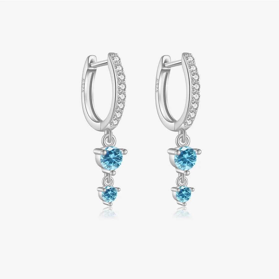 A pair of silver huggie hoop earrings with a row of small clear stones along the front, each dangling a three-stone cluster of light blue gemstones on a light gray background.