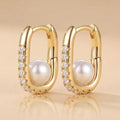 A pair of gold hoop earrings with a large white pearl and a row of clear cubic zirconia stones on the side, set against a warm beige background.