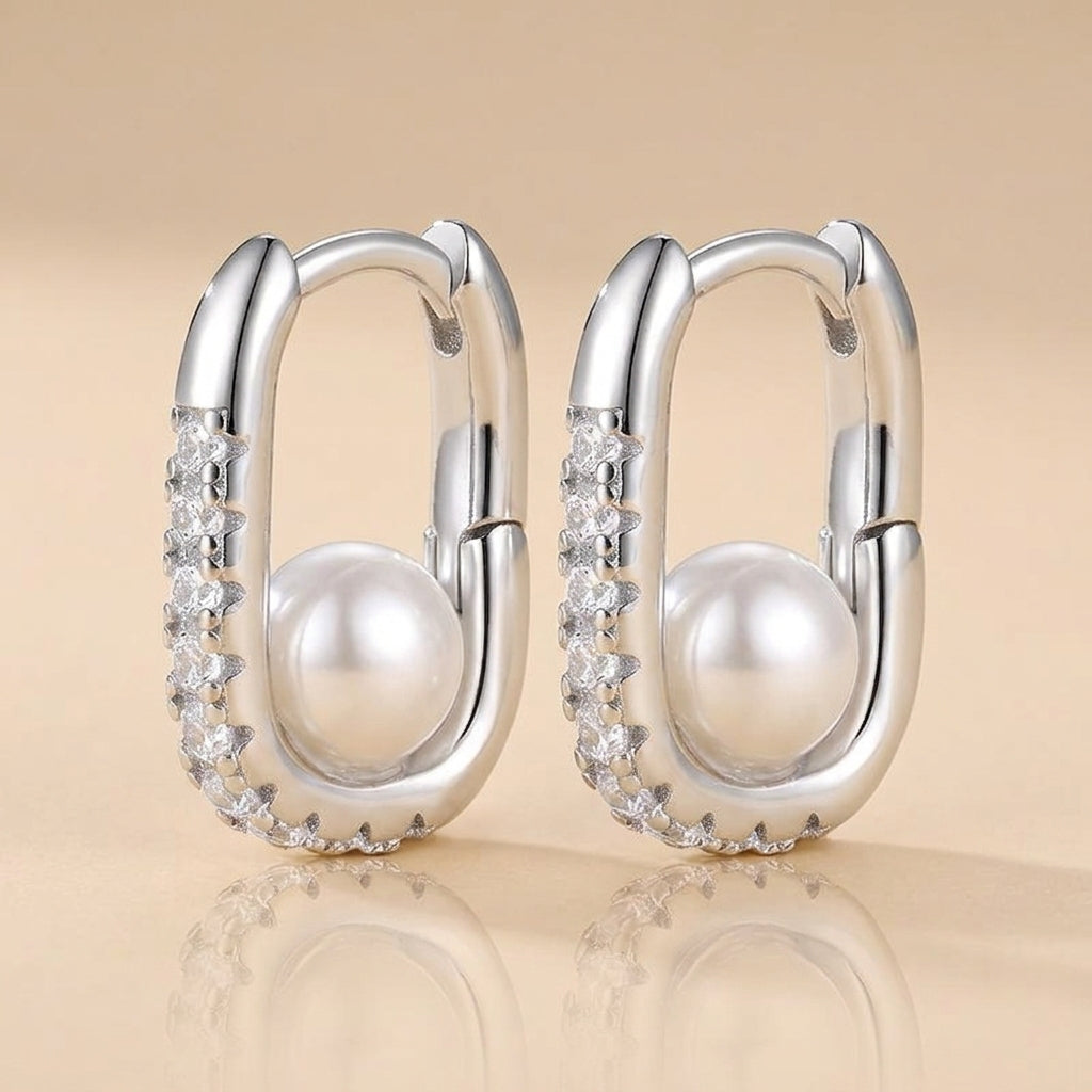 A pair of silver hoop-style earrings with large white pearls and rows of clear stones, shown on a warm beige reflective background.