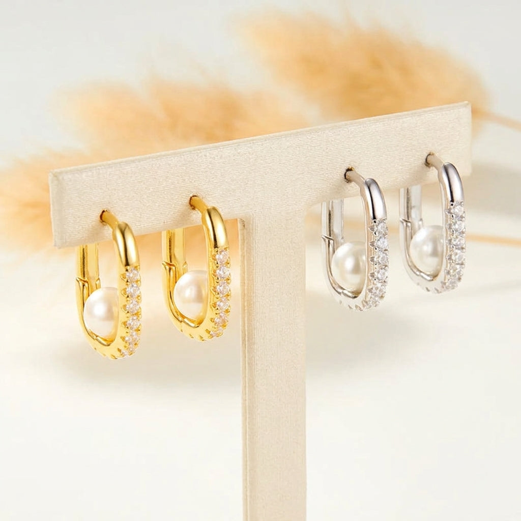 Two pairs of hoop earrings displayed on a cream jewelry stand. One pair is yellow gold with clear pavé stones and a white pearl, and the other pair is silver with clear pavé stones and a white pearl.