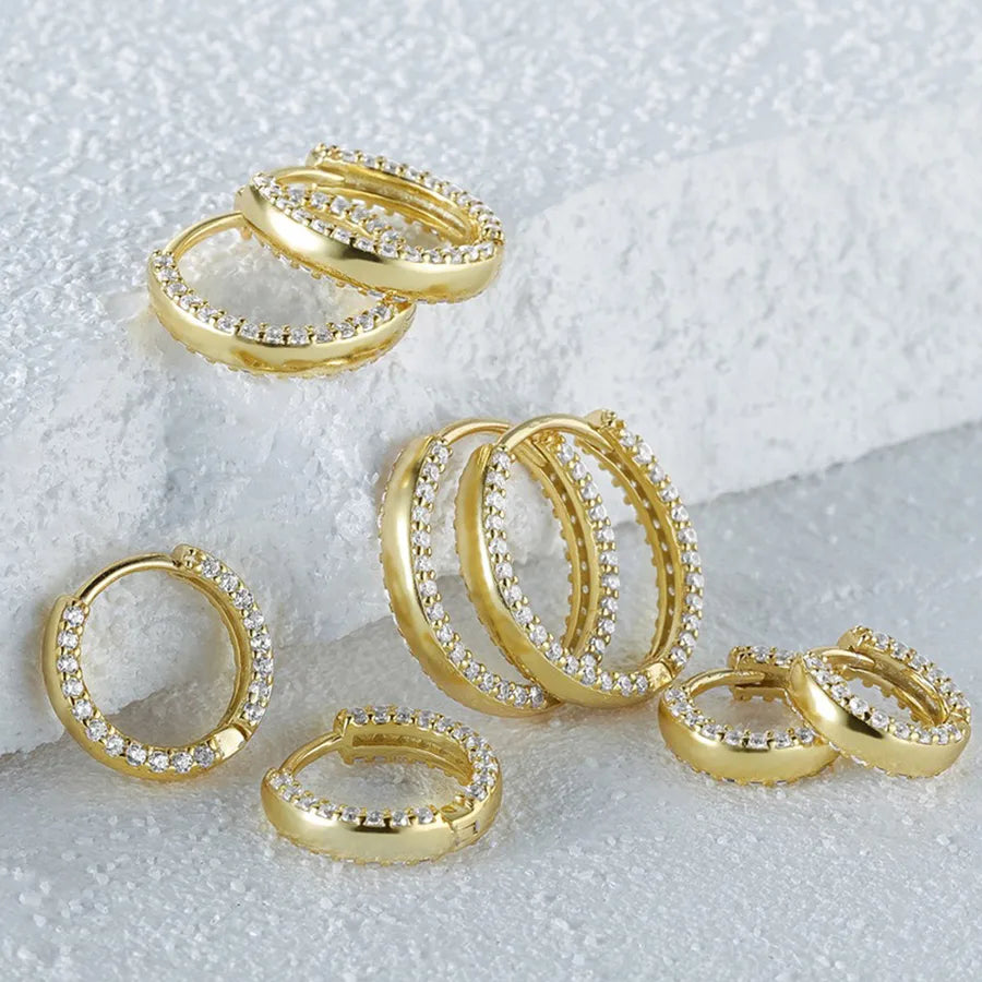 A set of gold huggie hoop earrings embellished with clear pavé stones, arranged on a textured white background.