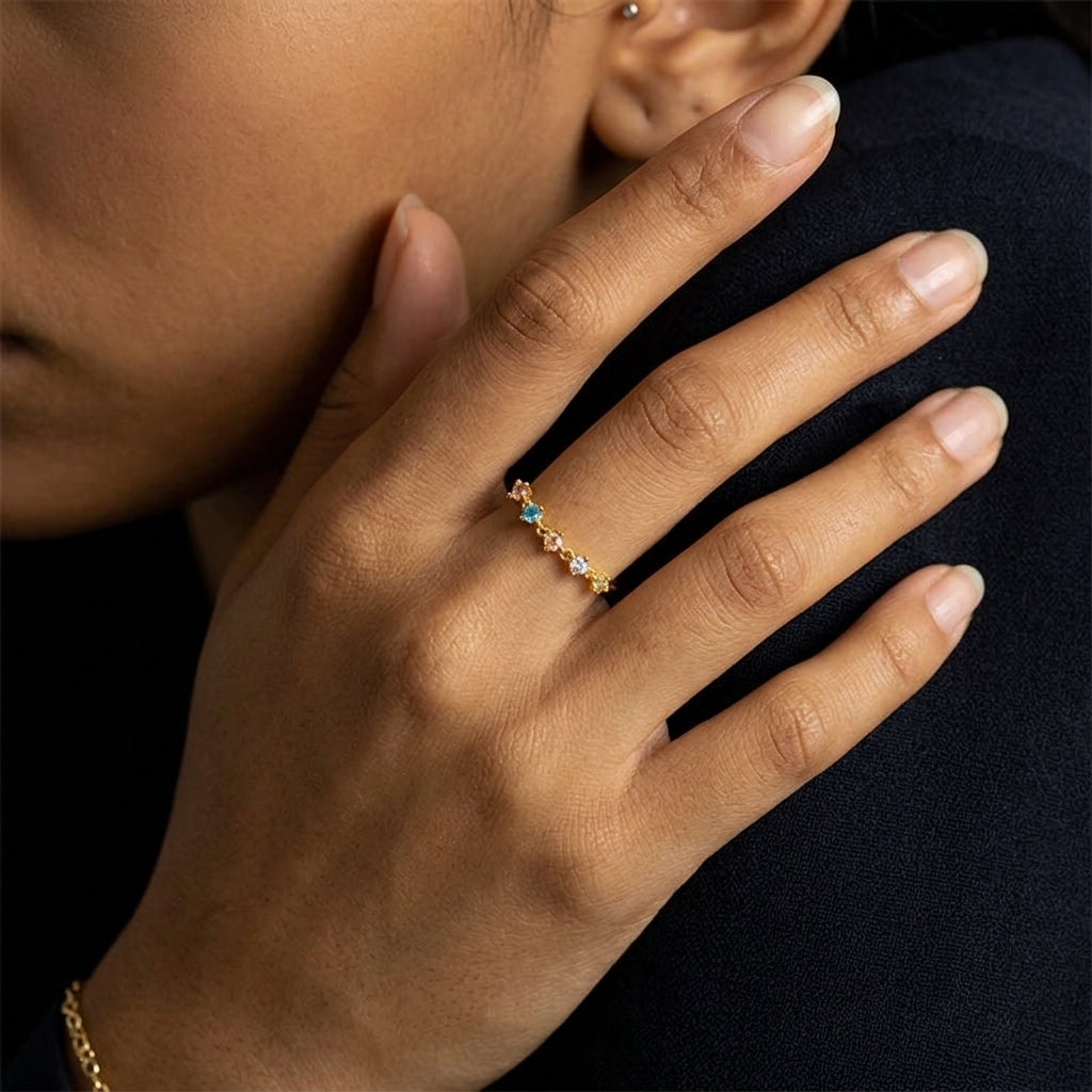 A close-up lifestyle shot of a model's hand wearing a delicate gold ring adorned with small colorful gemstones, set against a dark navy blue top.