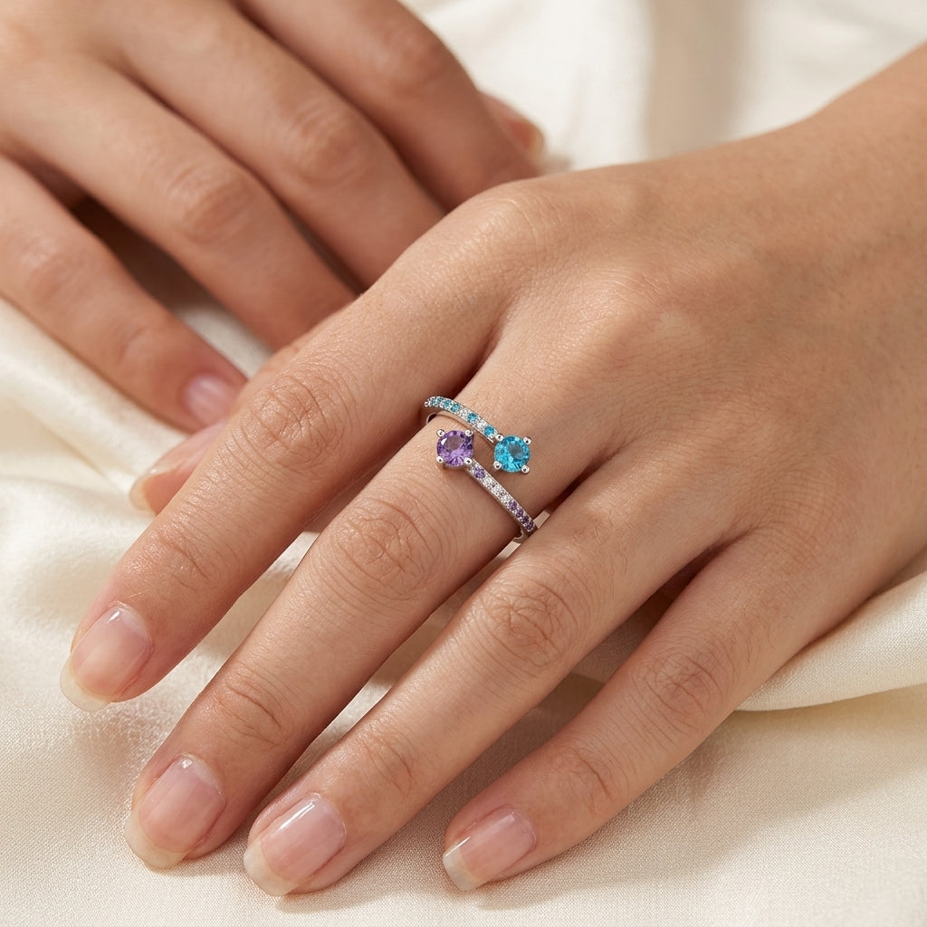 A close-up of a hand wearing a delicate silver ring with two heart-shaped gemstones, one light blue and one lavender purple, accented by small clear stones on a soft cream satin background.